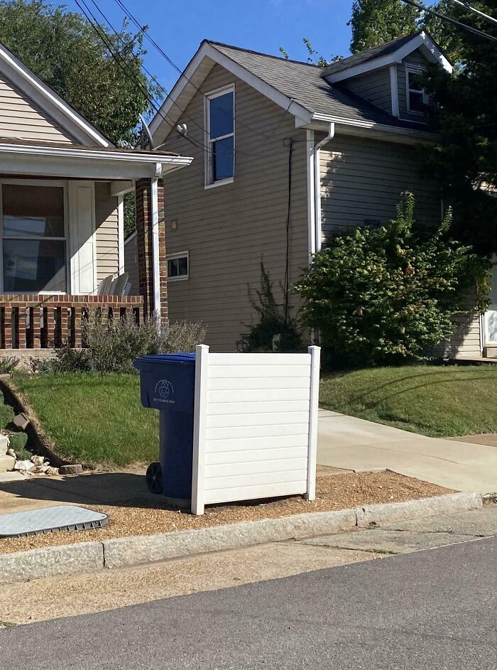 Blue recycling bin partially enclosed by a small DIY white fence on a sidewalk next to a house.