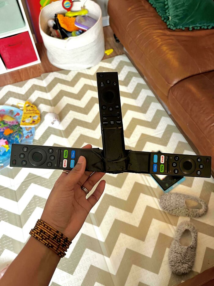 Hand holding a deeply cursed DIY project with three remote controls taped together in a living room setting.
