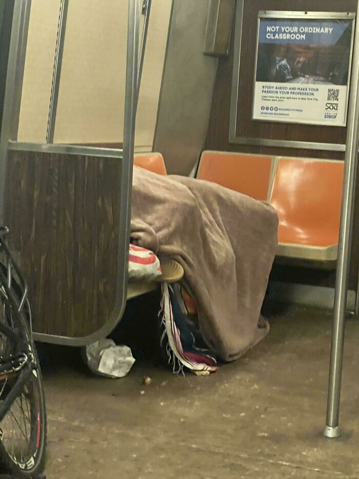 Person covered with a blanket lying on the floor of a NYC subway car next to empty orange seats and a bicycle.