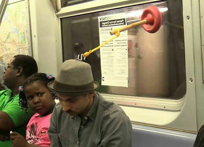 Man sitting on NYC subway with a plunger stuck to the window behind him in a weird subway moment.