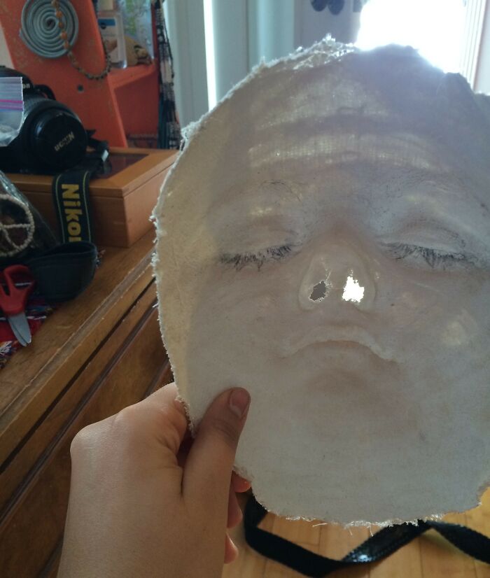 Hand holding a funny face mask with closed eyes and a distorted expression in a cluttered room, funny photos.