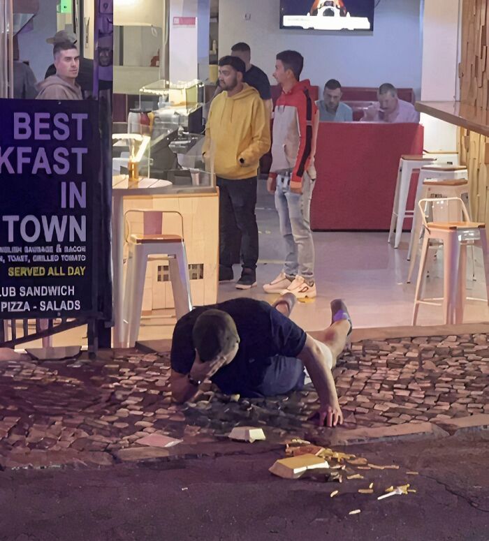 Man lying on cobblestone street with spilled food, while others stand near a restaurant in a funny photos moment.
