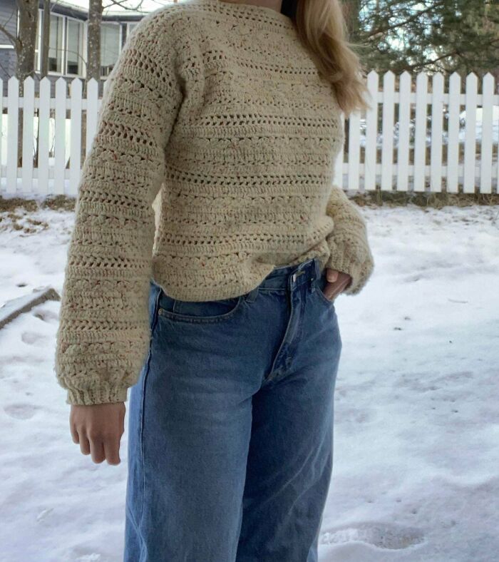 A person in a beautifully crocheted cream sweater and jeans, standing in the snow. An awesome crocheted item.