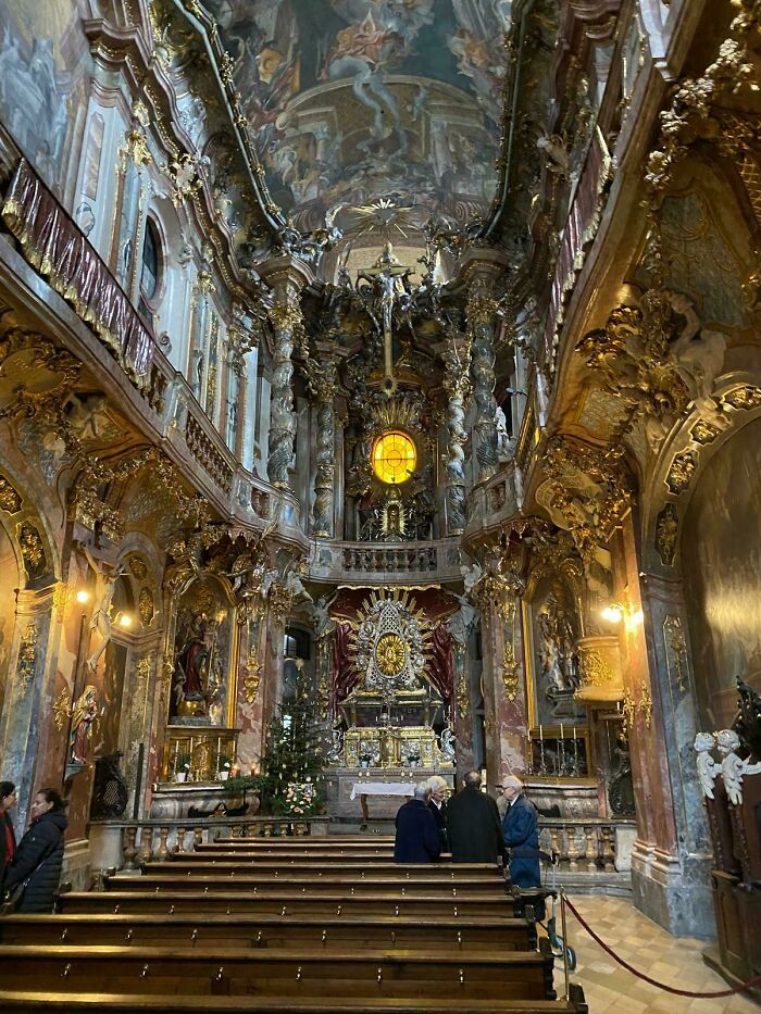 Ornate and beautiful architecture of a church interior with golden decorations, paintings, and people seated in pews.