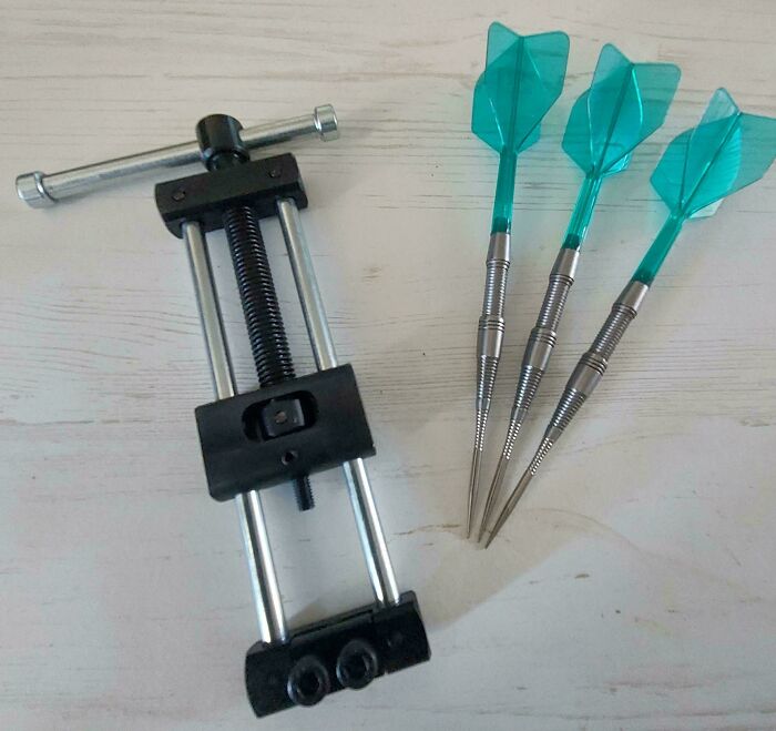 Tool clamp and three darts with teal flights on a light wooden surface, illustrating interesting tools solving small problems.