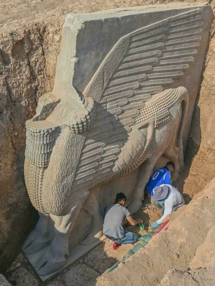 Ancient winged bull statue being excavated by workers, showcasing almost perfectly preserved antique items from history.