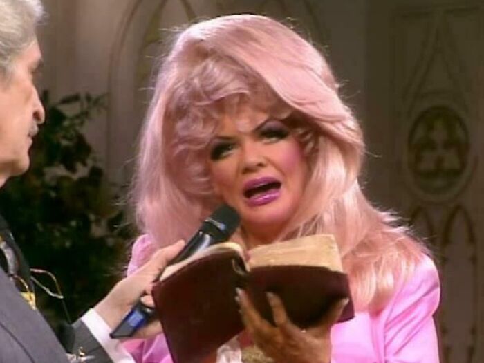 Woman with an exaggerated pink hairstyle and heavy makeup speaking into a microphone, showing tragic hair accident.