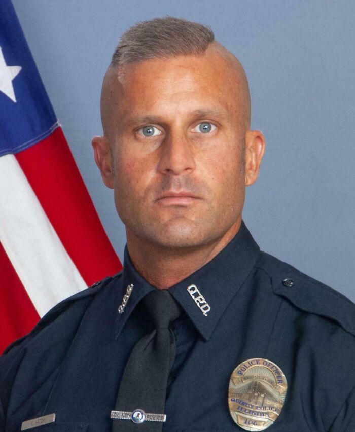 Police officer with a flawed haircut standing in front of an American flag, showcasing a tragic hair accident.