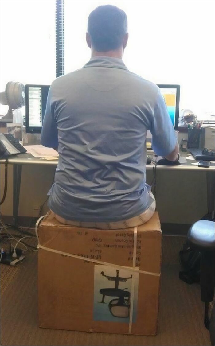 Man sitting on a cardboard box at a desk working on two computer screens, showing extreme laziness at the office.