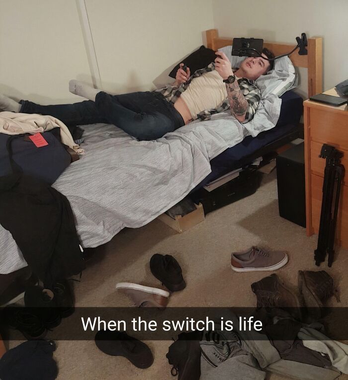 Young man showing extreme laziness using a phone mount to play Nintendo Switch while lying on a messy bed.