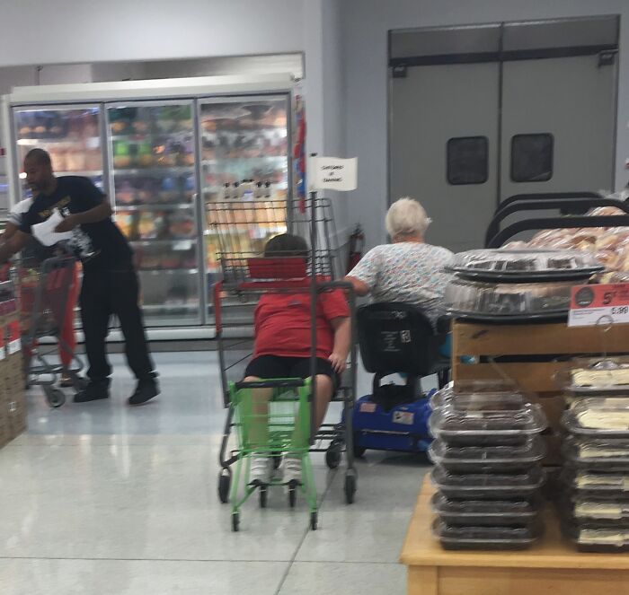 Person sitting lazily in a grocery cart while another uses a mobility scooter inside a store, showing extreme laziness.