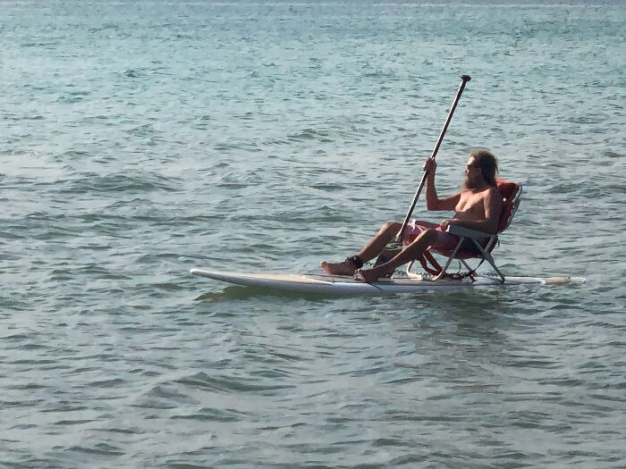 Man demonstrating extreme laziness by sitting on a chair while paddleboarding on calm water, embodying lazy lifestyle goals.