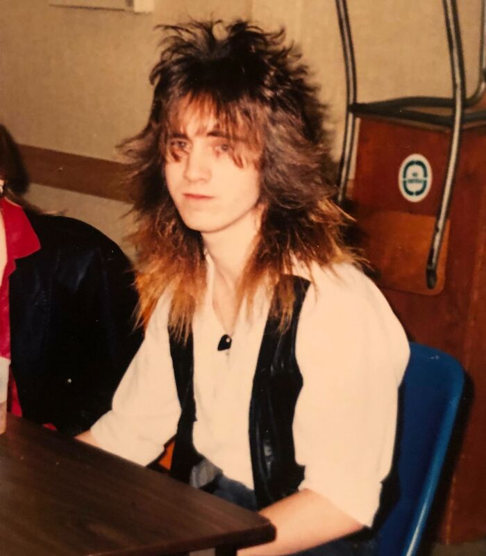Young person with an iconic 80s hairdo featuring voluminous layered spikes, wearing a leather vest and white shirt indoors.
