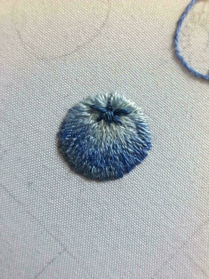 A close-up of an embroidery creation featuring a small blue and white shaded pom-pom stitch.