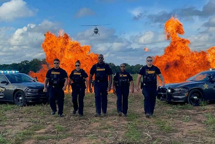 Group of sheriff officers walking in front of police cars with large fire explosions and a helicopter in the background