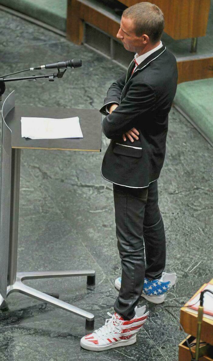Man in formal suit wearing cringe sneakers with wings and stars while standing at a podium giving a speech.