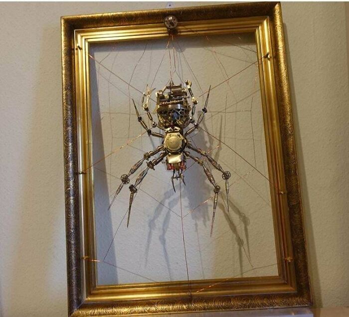 Metalworker-crafted spider sculpture suspended in a gold frame, showcasing intricate metalwork defying physics and logic.