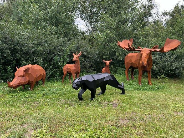 Geometric metal animal sculptures including a moose, bear, deer, and boar displayed outdoors on grass near trees.