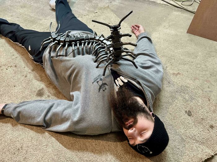 Man lying on the floor with a remarkable metal centipede sculpture placed on his chest showcasing metalworkers' creativity.