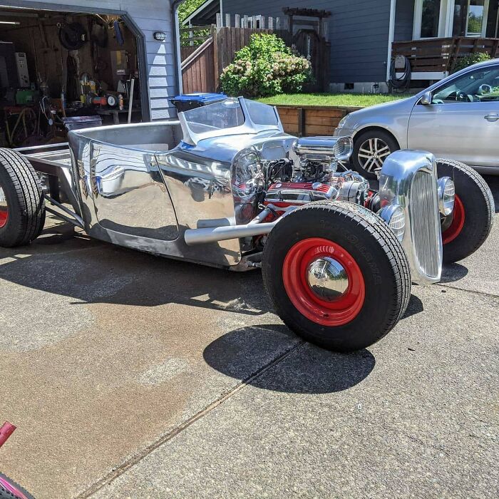 Custom polished metal hot rod with exposed engine and red wheels parked outside a garage showcasing metalworkers' craftsmanship.