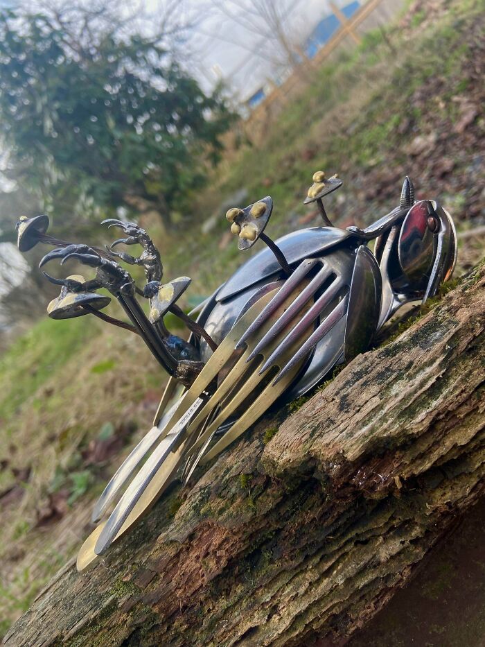 Metalworker sculpture of a bird made from forks and spoons resting on a mossy log outdoors.
