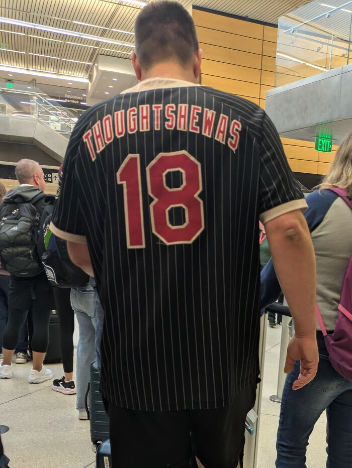 Man wearing a black pinstripe jersey with the phrase "THOUGHTSHEWAS 18" showcasing a cringe moment in a busy indoor setting.
