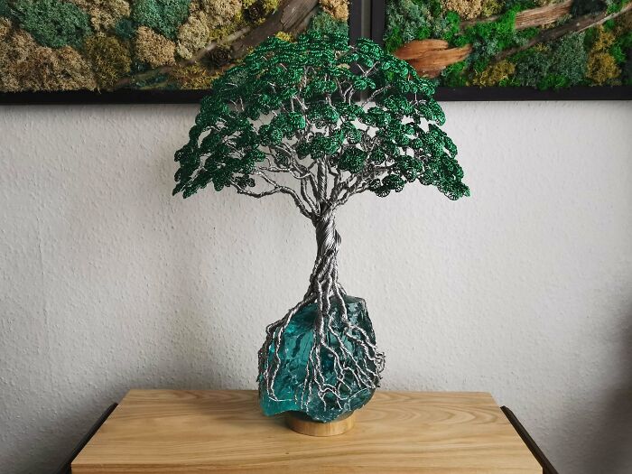 Intricately crafted metalworkers sculpture of a tree with detailed roots and green wire leaves on a rock base.