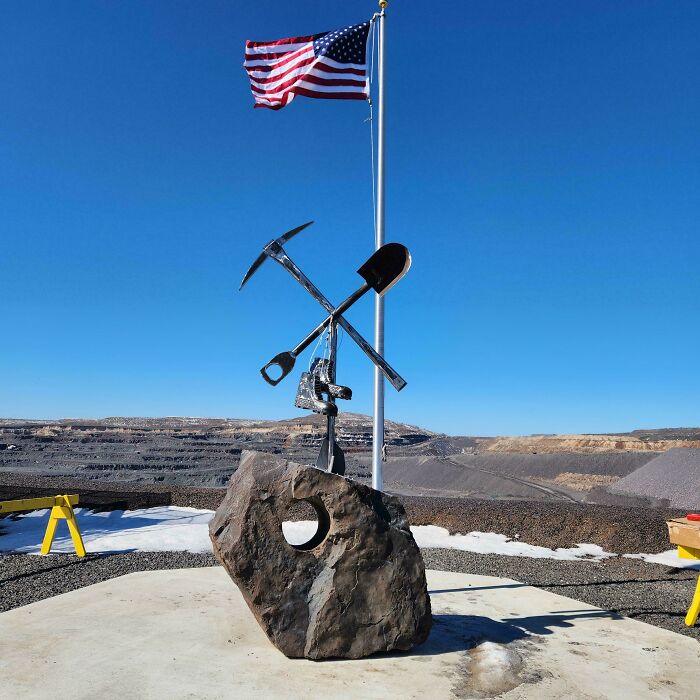 Metalworkers sculpture with pickaxe and shovel crossed on rock base near American flag under clear blue sky.