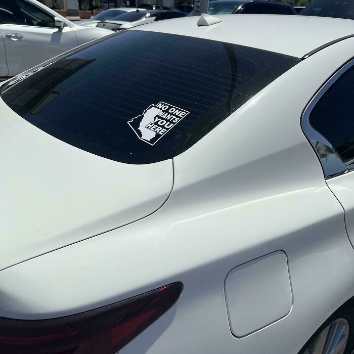 White car with a sticker on the rear window saying no one wants you here, showing a cringe moment captured in public.
