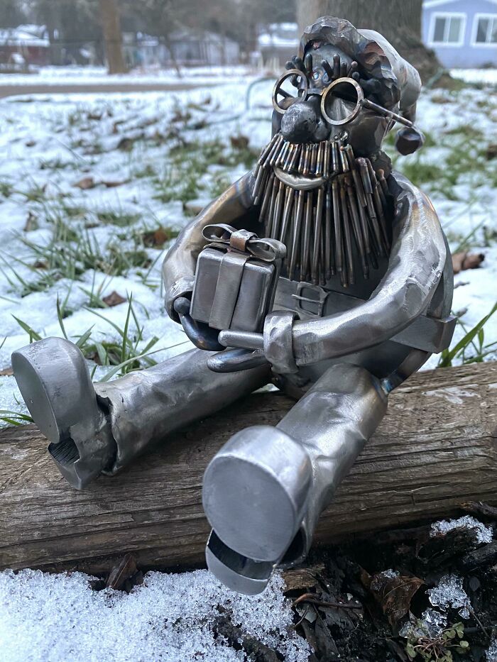 Metalworker sculpture of a bearded figure wearing glasses, holding a gift, sitting on a log in snowy outdoor setting.