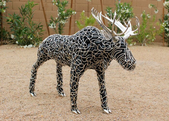 Metalworker sculpture of a moose made from intertwined metal rings standing outdoors on gravel near plants.