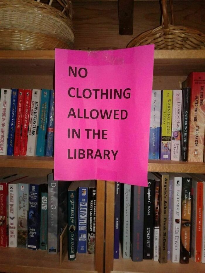 Bright pink sign reading no clothing allowed in the library displayed on a bookshelf with various books, featuring strange signs.