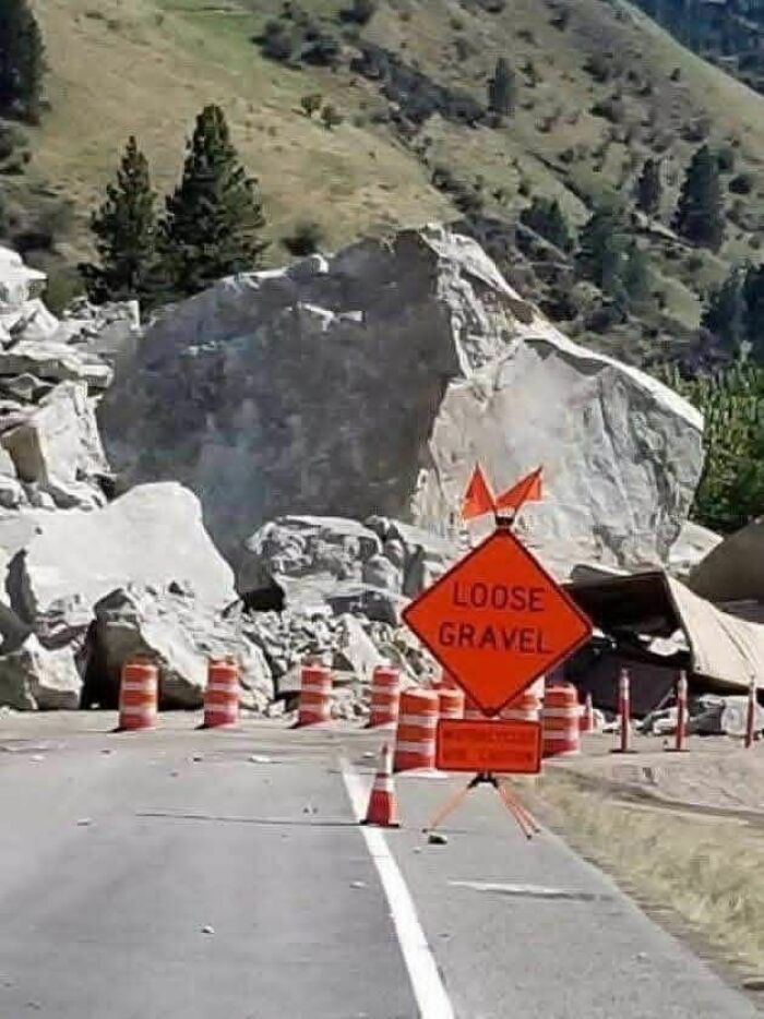 Road with an orange loose gravel sign and traffic cones in front of a large boulder blocking the highway strange signs.