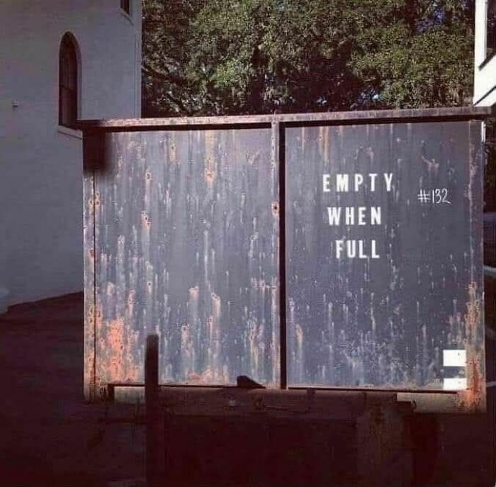 Rusty metal sign on a gate displaying the strange message empty when full in an outdoor setting with trees and a building nearby