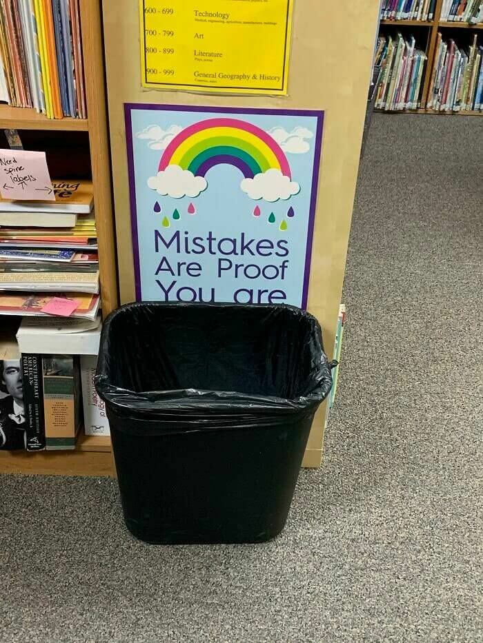 Library scene with a colorful rainbow sign about mistakes placed oddly behind a black trash bin, showing strange signs to double take.