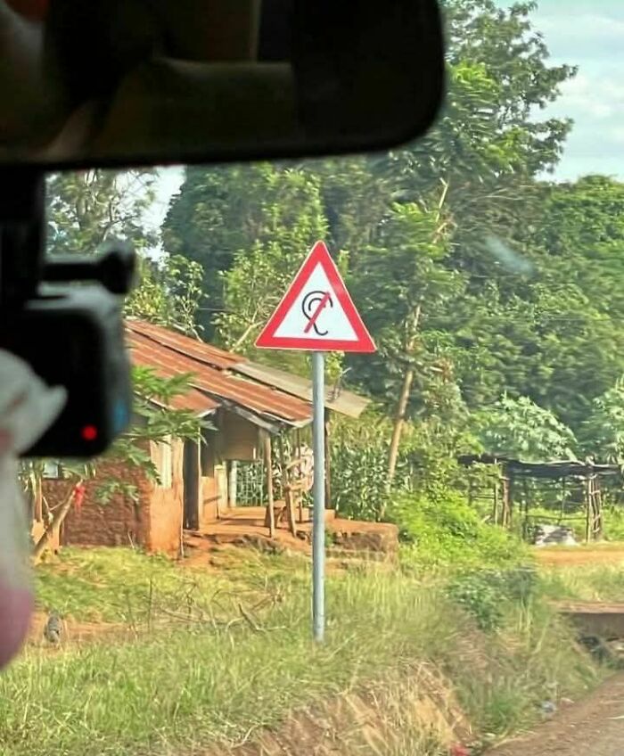 Unusual strange road sign with a distorted symbol on a rural roadside surrounded by greenery and houses.