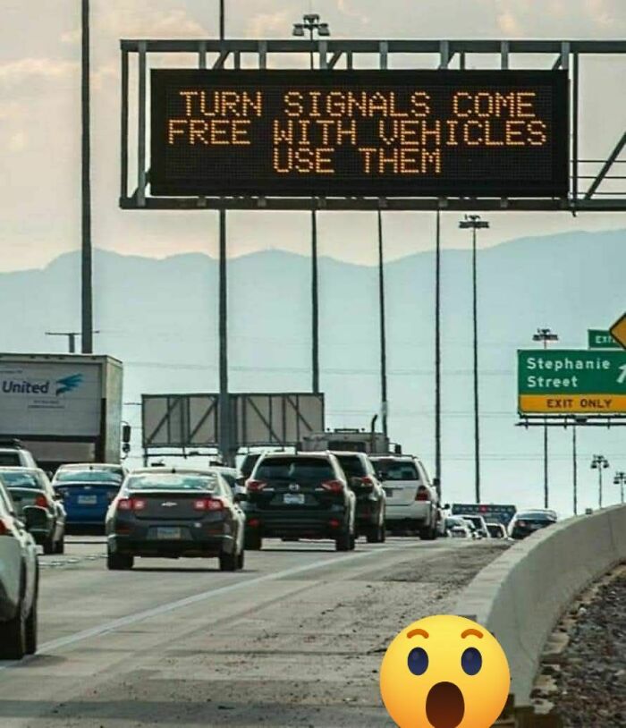 Highway traffic under a strange electronic sign humorously urging drivers to use free turn signals with vehicles.
