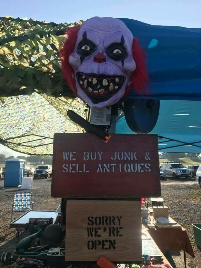 Creepy clown head above a sign selling antiques with a confusing apology sign at an outdoor market.