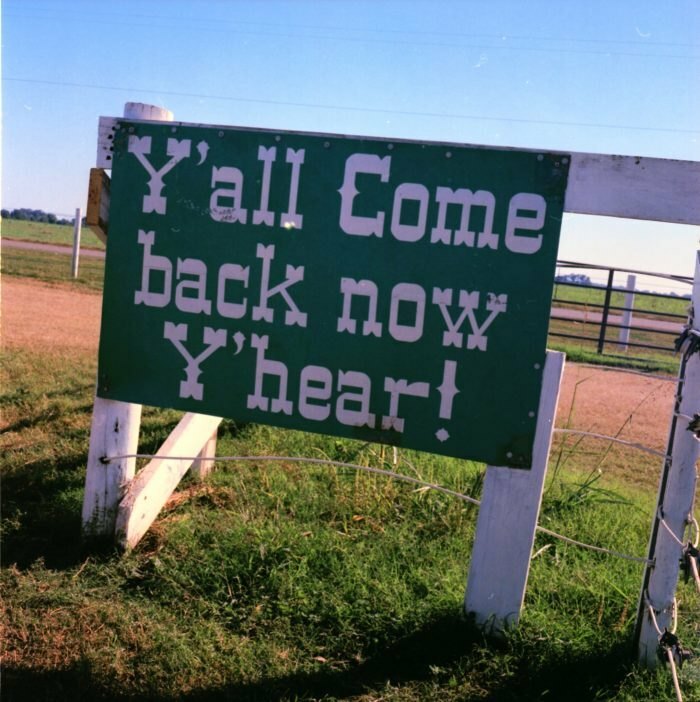 Green roadside sign with white text saying Y'all Come back now Y'hear in a rural outdoor setting, a strange sign example.