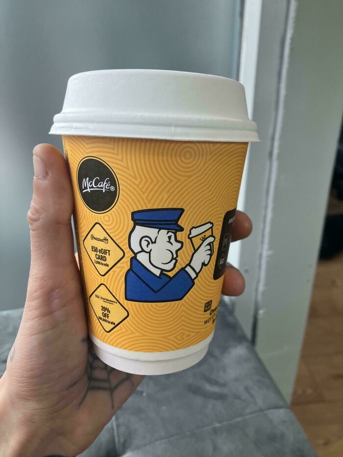 Hand holding a McCafé coffee cup with a confusing design showing a man holding a tiny coffee cup illustration.