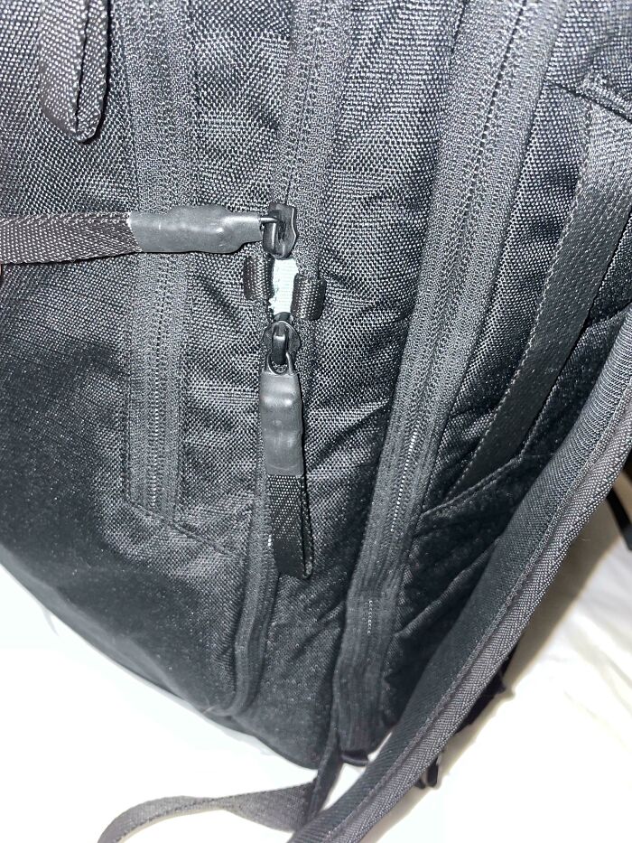 Close-up of a black backpack with defective zipper design showing zipper pullers that cannot properly close.
