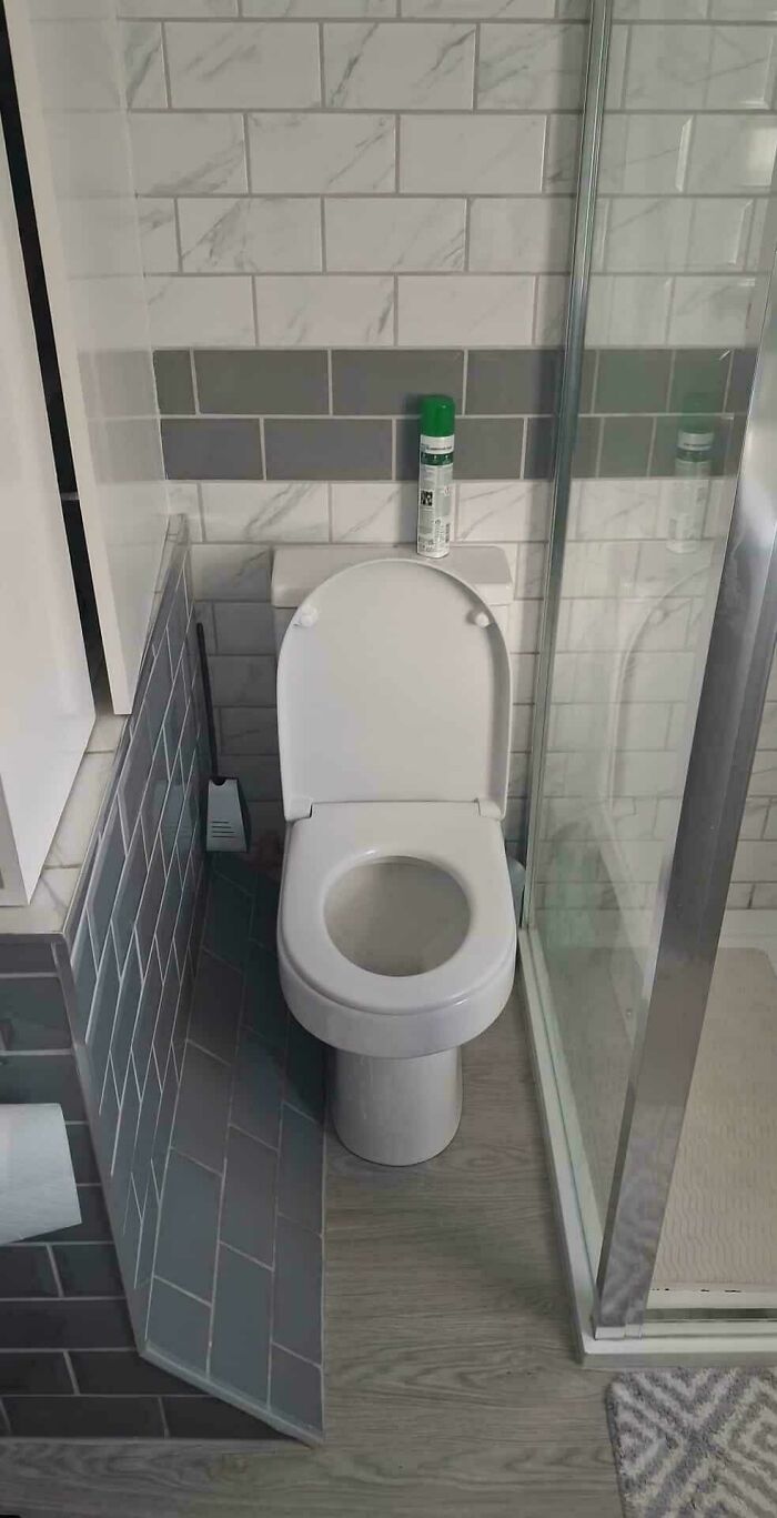 Toilet installed in a cramped bathroom with awkward angled wall and shower enclosure, showcasing terrible product design.