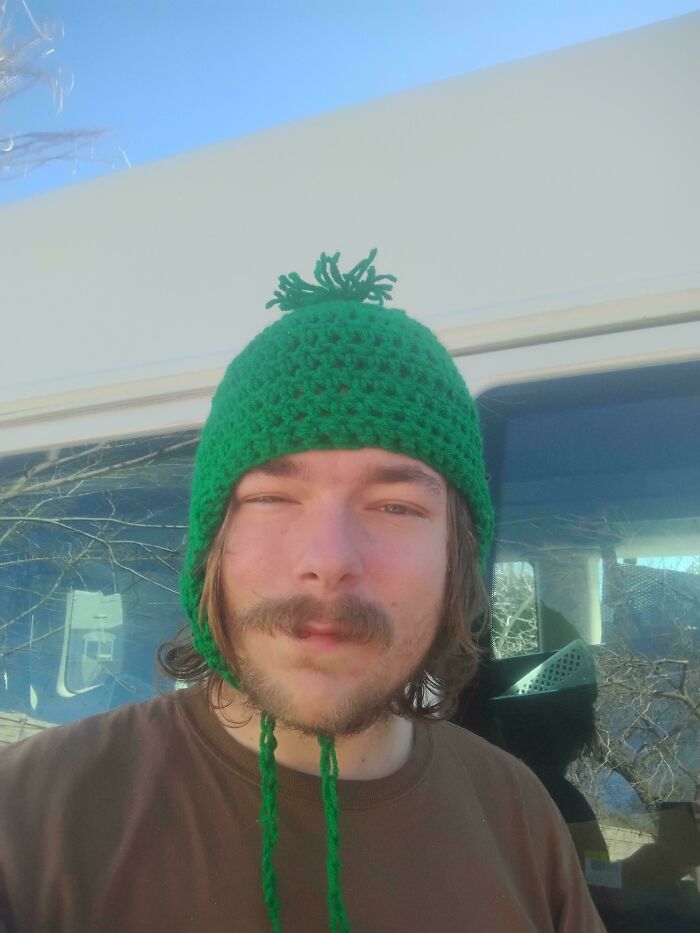A person wearing a green knitted beanie with earflaps and a pom-pom, a cool find from dumpster diving.