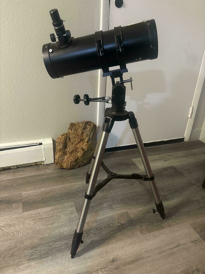 A black telescope on a tripod standing on a wooden floor, a fantastic item discovered through dumpster diving.