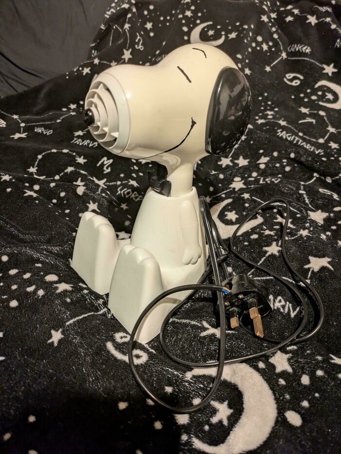 A Snoopy-shaped Buy It For Life fan on a black blanket with constellations. The vintage fan has an electrical cord.