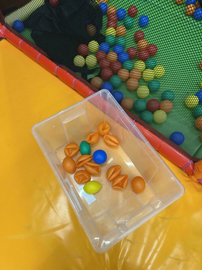 A clear bin with some deflated orange and colorful balls sits on a yellow mat, next to a ball pit, offering mildly interesting pictures.