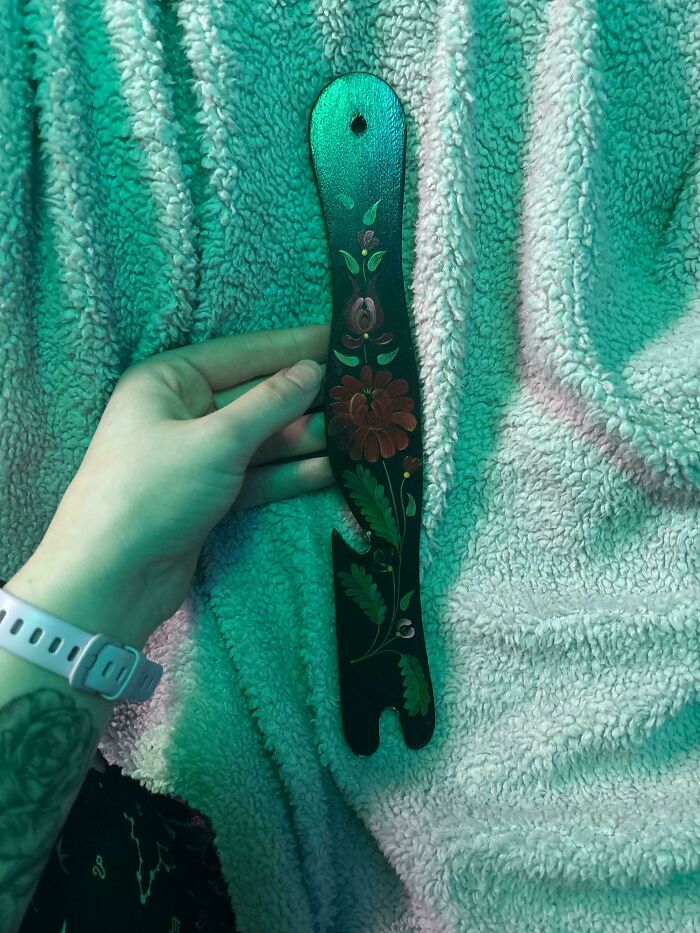 Hand holding a strange floral-painted wooden object with an unusual shape on a textured blanket.