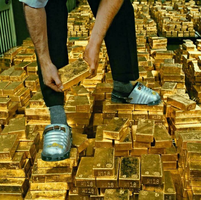 Person standing on stacks of gold bars wearing silver shoes, holding a gold bar, a strange pic causing confusion