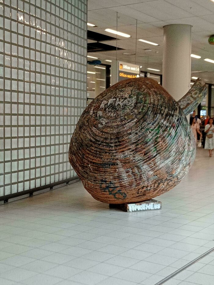 Large wooden sculpture resembling a giant acorn covered in graffiti, displayed indoors in a public space, strange pics keyword.