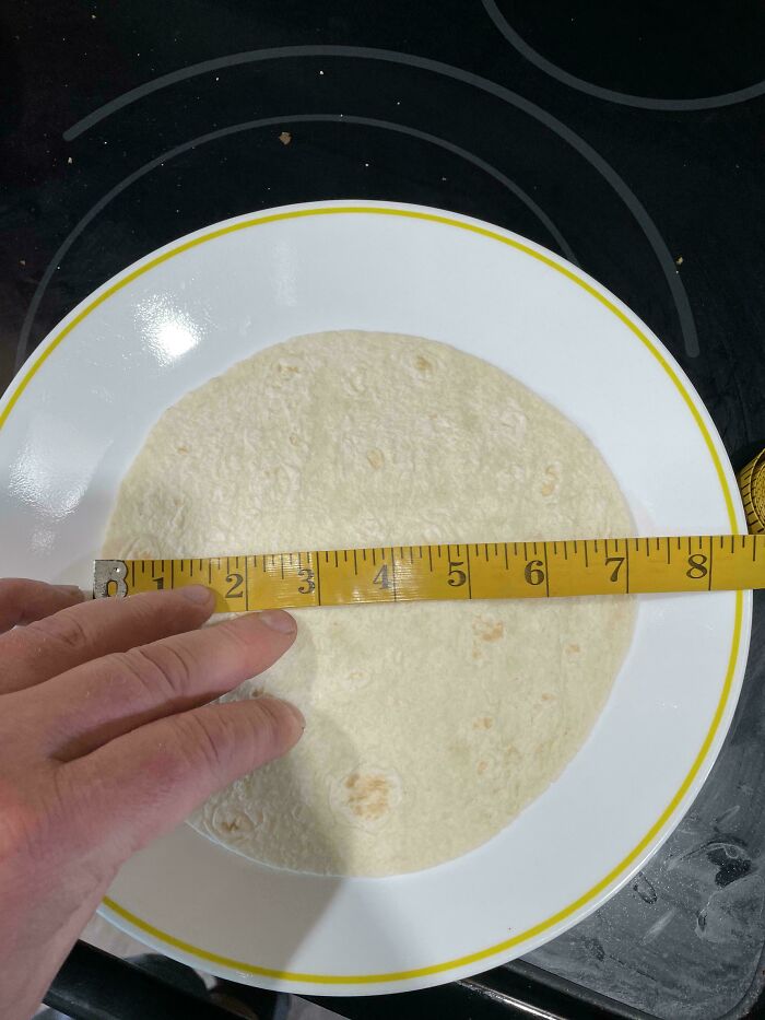 Hand measuring a small tortilla on a plate, illustrating shrinkflation in American products with a tape measure.